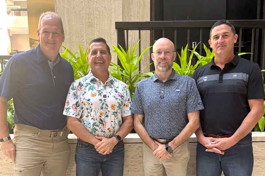 Four men smiling, standing in front of green plants at a leadership offsite.