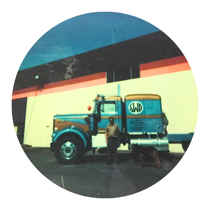 Man standing beside blue and brown truck with "SWD" logo, industrial building in background