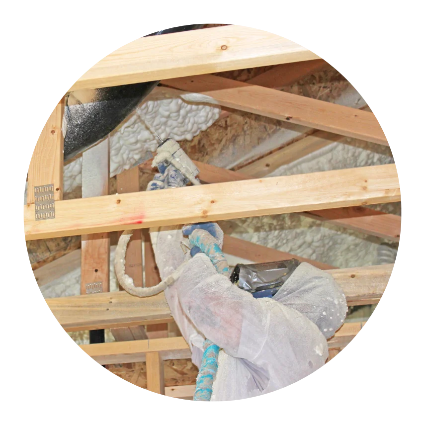 Worker in protective gear applying spray foam insulation to a wooden attic ceiling
