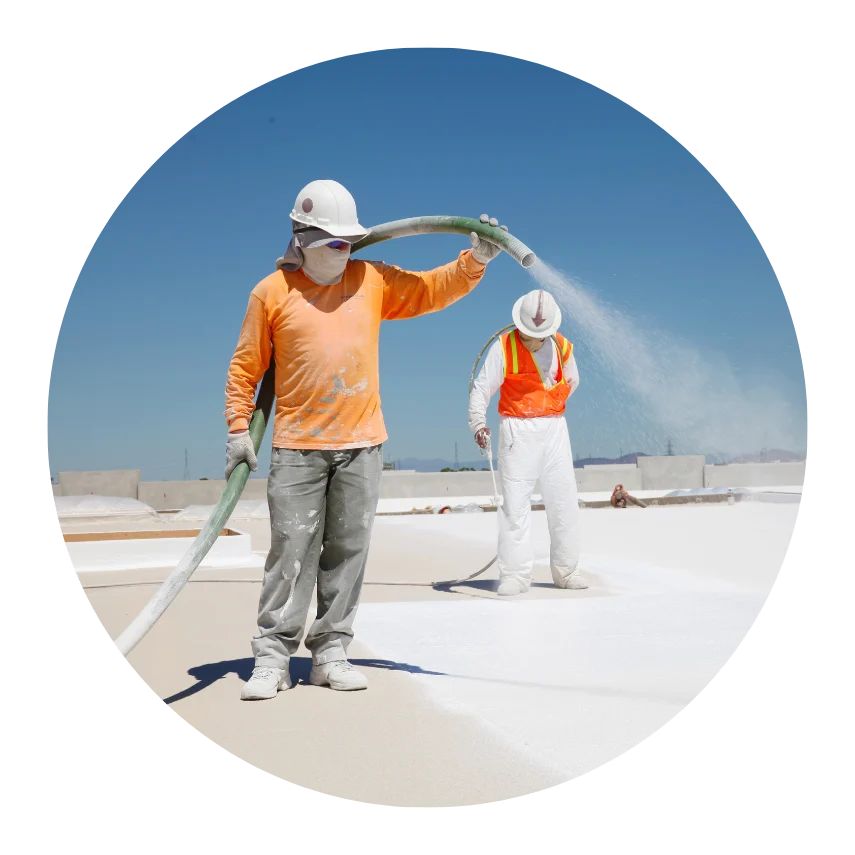 Construction worker in safety gear spraying spray foam roofing insulation on a flat roof under a clear blue sky.