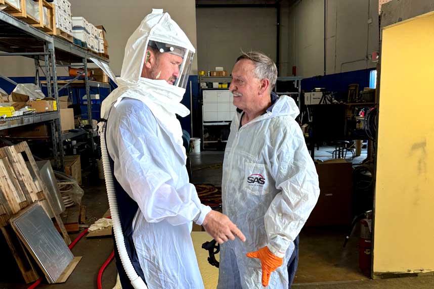 Two team members in protective suits, one wearing a full-face respirator, preparing for spray foam insulation work inside a warehouse.